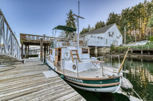 A boat docked at a wooden pier, surrounded by trees and a white building in the background.