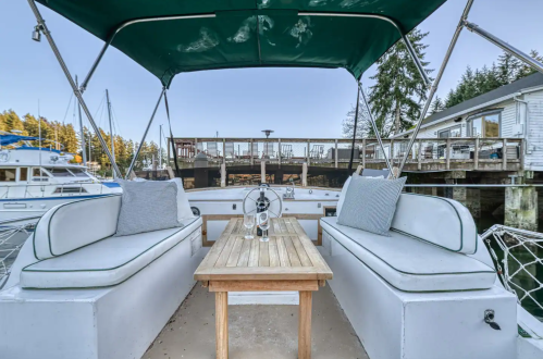 A boat's deck with cushioned seating and a wooden table, overlooking a marina with nearby docks and trees.