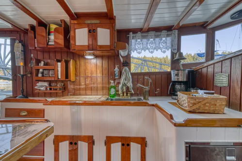 Cozy boat interior featuring a wooden kitchen with a sink, coffee maker, and decorative shelves. Natural light filters in.