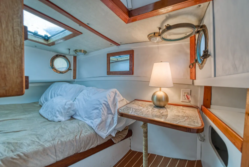Cozy cabin interior with a bed, lamp, and nautical decor, featuring porthole windows and wooden accents.
