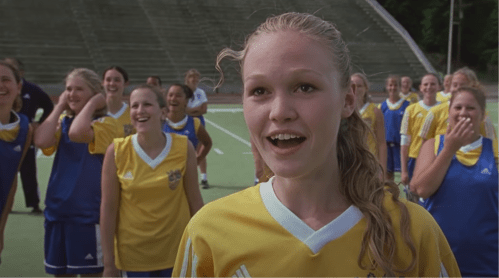 A young female soccer player smiles excitedly, surrounded by teammates in yellow and blue jerseys, celebrating on a field.