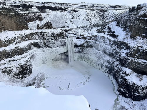 A frozen waterfall cascades into a snowy canyon, surrounded by icy cliffs and a winter landscape.