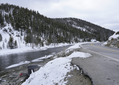 A snowy landscape with a winding road beside a river, surrounded by evergreen trees and mountains in the background.