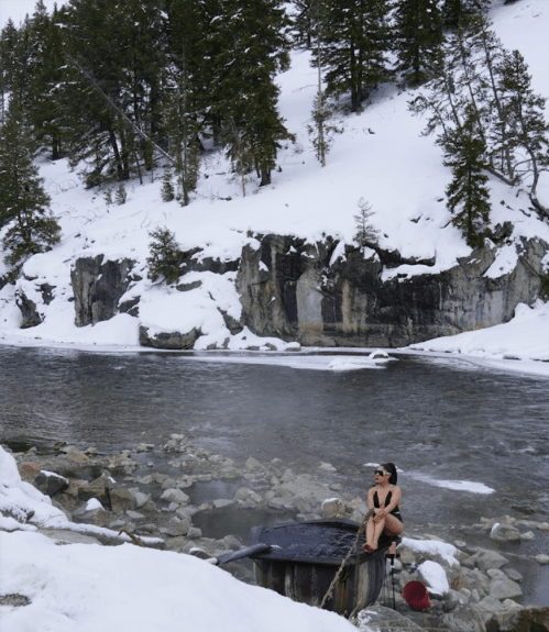 A person sits by a steaming hot spring near a snowy river, surrounded by rocky cliffs and evergreen trees.