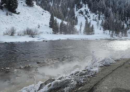 A snowy landscape with a river, steam rising from the water, and pine trees lining the banks.