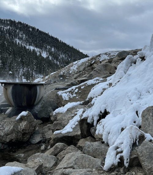 A metal pot sits on rocky terrain, surrounded by snow and ice, with a mountainous landscape in the background.