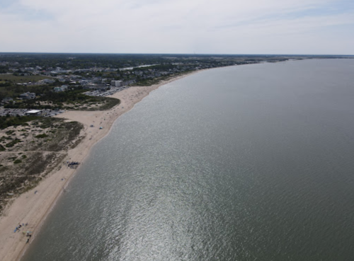 Aerial view of a sandy beach curving along a calm sea, with distant shoreline and scattered beachgoers.