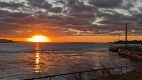 A vibrant sunset over a calm waterway, with clouds reflecting orange and gold hues, and a pier in the foreground.