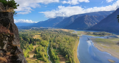 A scenic view of a river winding through lush green valleys and mountains under a blue sky with scattered clouds.