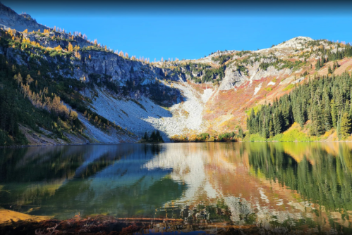 A serene mountain lake reflecting colorful autumn foliage and rocky cliffs under a clear blue sky.
