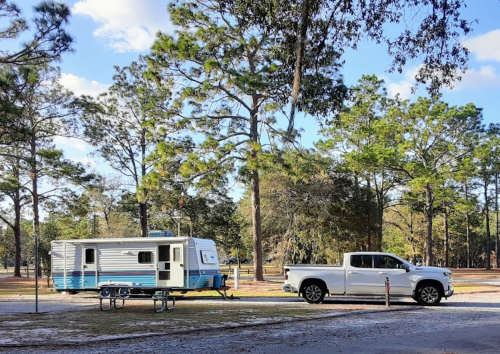 A white pickup truck towing a blue and white camper trailer in a wooded park setting.