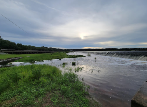 A serene landscape featuring a river with a waterfall, lush green grass, and a cloudy sky at sunset.