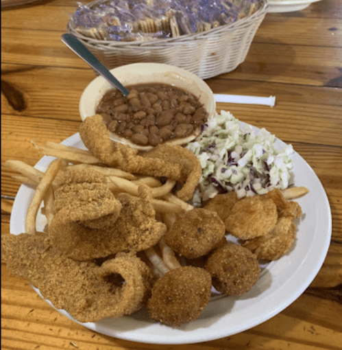 A plate of fried fish, hushpuppies, coleslaw, French fries, and a bowl of baked beans on a wooden table.
