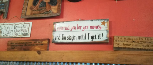 A rustic sign on a wall reads, "I married you for yer money... and I'm stayin' until I get it!"