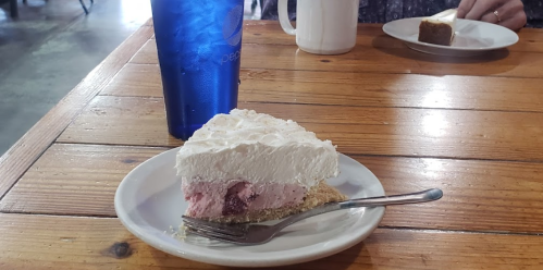 A slice of pink cream pie with whipped topping on a white plate, next to a blue glass and a piece of cake in the background.