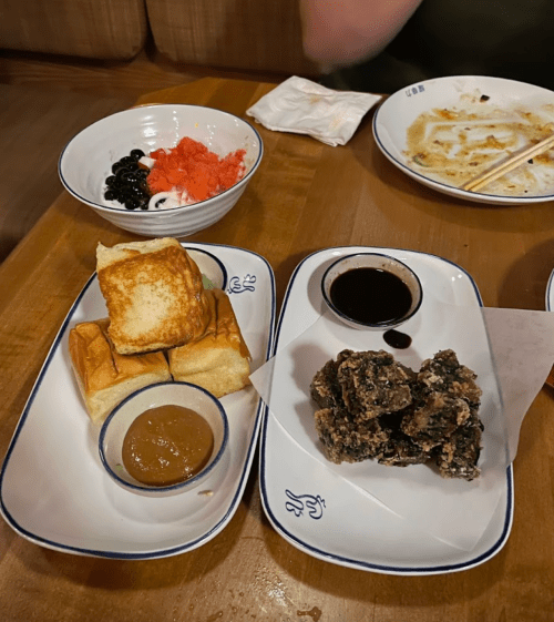 A wooden table with two plates: one with toast, sauce, and a green dish; the other with a bowl of colorful toppings.