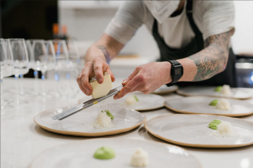 A chef garnishes plates with a grater, preparing a dish in a modern kitchen with wine glasses in the background.