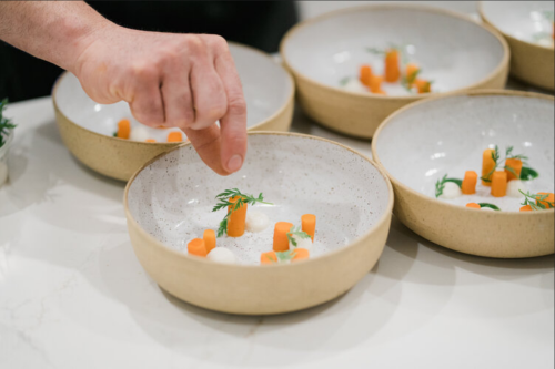 A hand sprinkles seasoning over a dish with carrots and creamy elements in a shallow bowl.