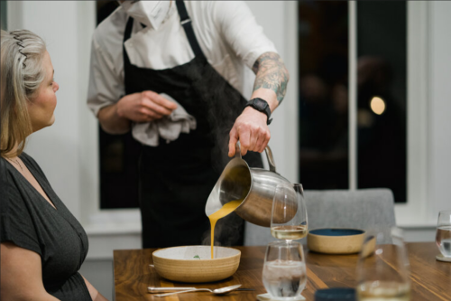 A chef pours soup into a bowl while a woman watches at a dining table, with glasses of wine nearby.