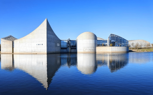 Modern architectural building with unique shapes, reflecting in calm water under a clear blue sky.