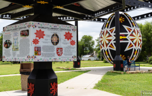 A large, colorful egg sculpture stands beside an informational sign about the world's largest Czech egg in a park.