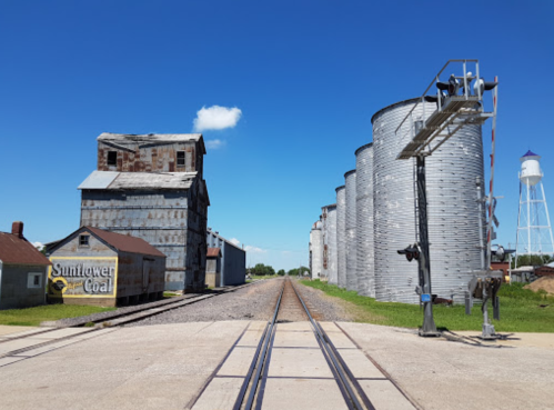 A rural scene featuring railroad tracks, silos, and old buildings under a clear blue sky.