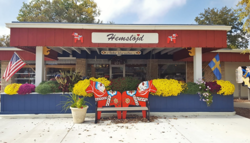 A colorful storefront named "Hemslöjd" with a Dala horse bench, flowers, and flags in a charming setting.