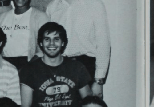 A black and white photo of a group of smiling individuals, with a young man in a T-shirt sitting in the front.