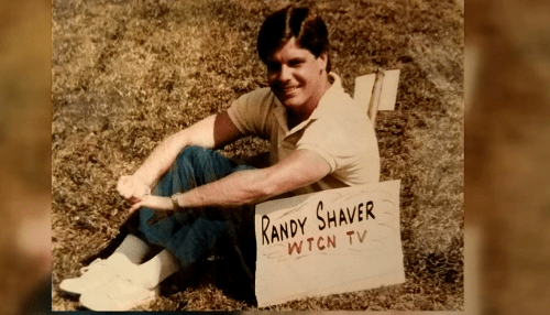A smiling man sits on the grass next to a sign that reads "Randy Shaver WTCN TV."