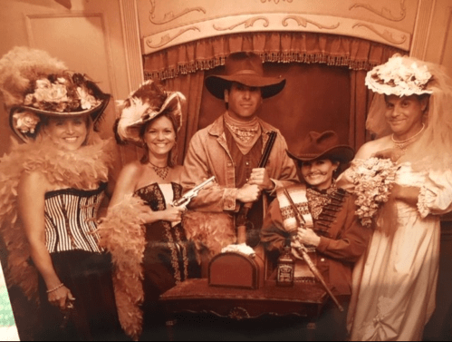 A sepia-toned photo of five people in Western attire, posing with props like guns and flowers in a vintage setting.