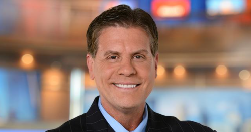 A smiling man in a dark suit and blue shirt stands in front of a blurred news studio background.