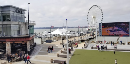 A waterfront scene featuring a ferris wheel, boats, and people enjoying a park area near a large screen.