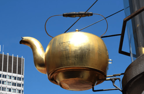 A large, shiny golden teapot sculpture hanging against a blue sky, with a label indicating its capacity.