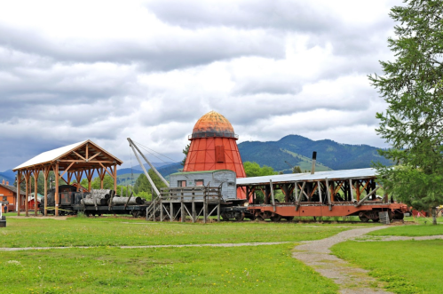 Historic train and wooden structures in a grassy area, with mountains and cloudy skies in the background.