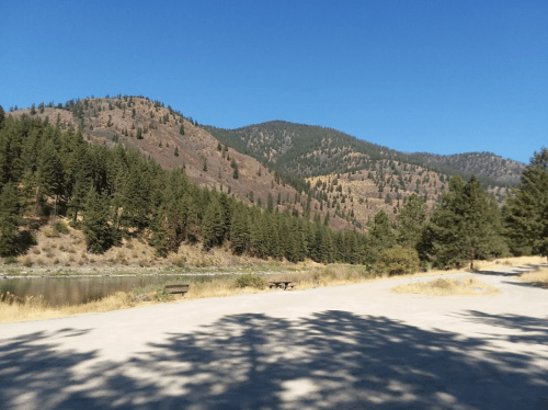 A scenic view of mountains and trees beside a calm river under a clear blue sky.