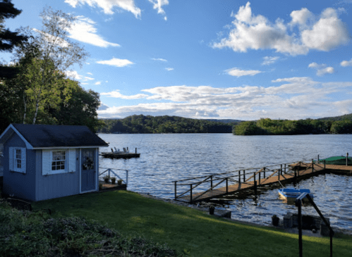 A serene lakeside view featuring a small blue shed, a dock, and boats under a partly cloudy sky.