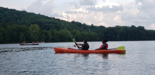 Two people kayak on a calm lake surrounded by green hills, with another kayak in the background.
