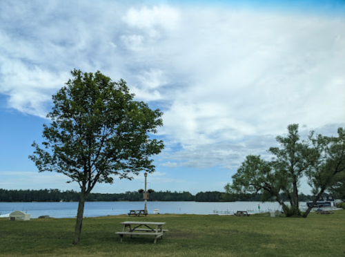 A serene lakeside view with trees, a picnic table, and a cloudy sky reflecting on the water.
