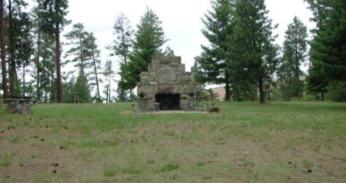 A stone fireplace surrounded by trees in a grassy area, with picnic tables nearby.