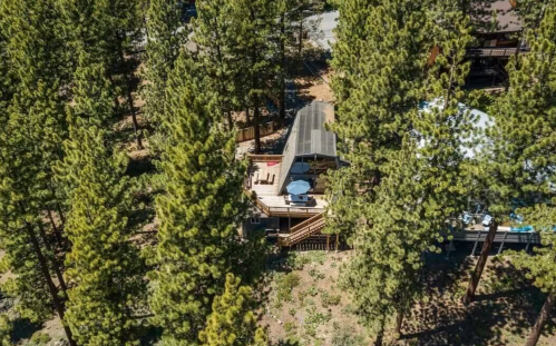 Aerial view of a cabin nestled among tall pine trees, featuring a deck with outdoor furniture.
