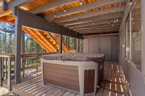 A wooden deck with a hot tub, surrounded by trees and a staircase leading to the upper level of a cabin.