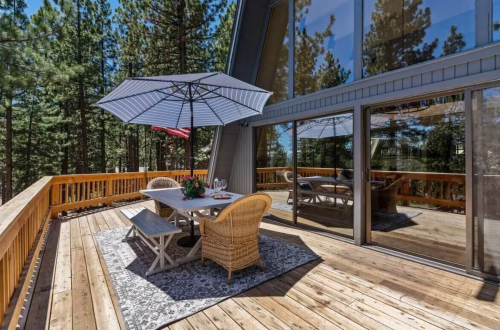 A wooden deck with a dining table, chairs, and an umbrella, surrounded by tall trees and large windows.