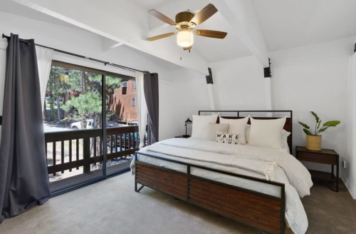 Bright bedroom with a large bed, ceiling fan, and sliding glass door leading to a balcony with greenery outside.