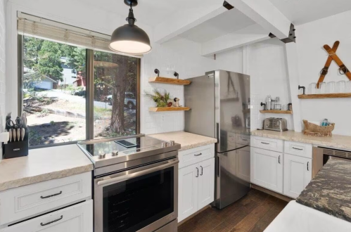 Modern kitchen with white cabinets, stainless steel appliances, and wooden shelves, featuring a large window with a view.