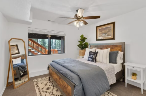 Cozy bedroom with a wooden bed, large window, and a mirror, featuring neutral decor and a warm atmosphere.