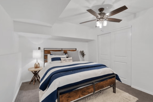 Cozy bedroom with a striped blue and white bedspread, ceiling fan, and a lamp on a wooden nightstand.