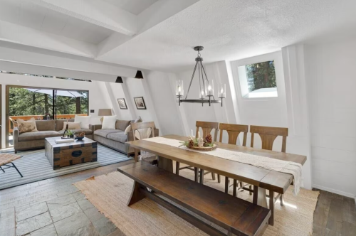 A modern dining area with a wooden table, chairs, and a cozy living space visible through large windows.