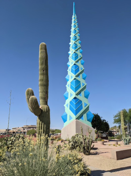 A tall, blue geometric sculpture stands beside a tall cactus in a desert landscape under a clear blue sky.