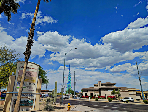 A vibrant sky with clouds above a street scene featuring palm trees, a pharmacy sign, and a colorful tower in the distance.