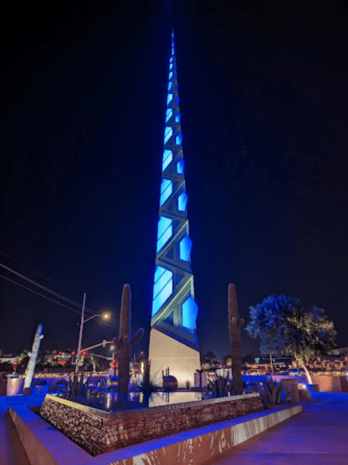 A tall, illuminated blue and green spire at night, surrounded by cacti and urban scenery.